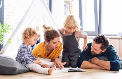 young family with two small children indoors in bedroom reading a book.