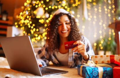smiling african american woman using laptop, sitting near christmas tree with card, shopping online